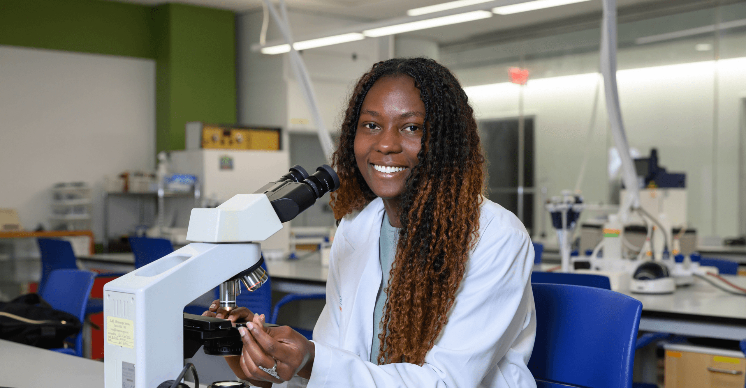 Smiling woman wearing a lab coat sits behind a microscope
